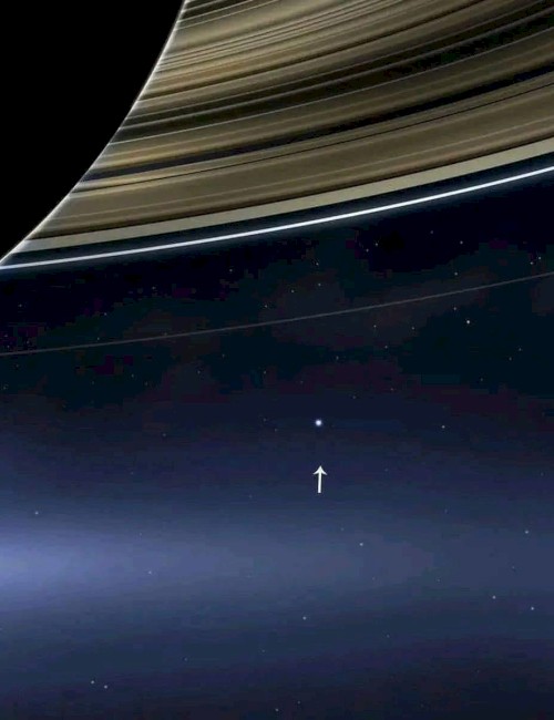The image shows Saturn's rings with a small dot in the distance, highlighted by an arrow, representing Earth from afar.